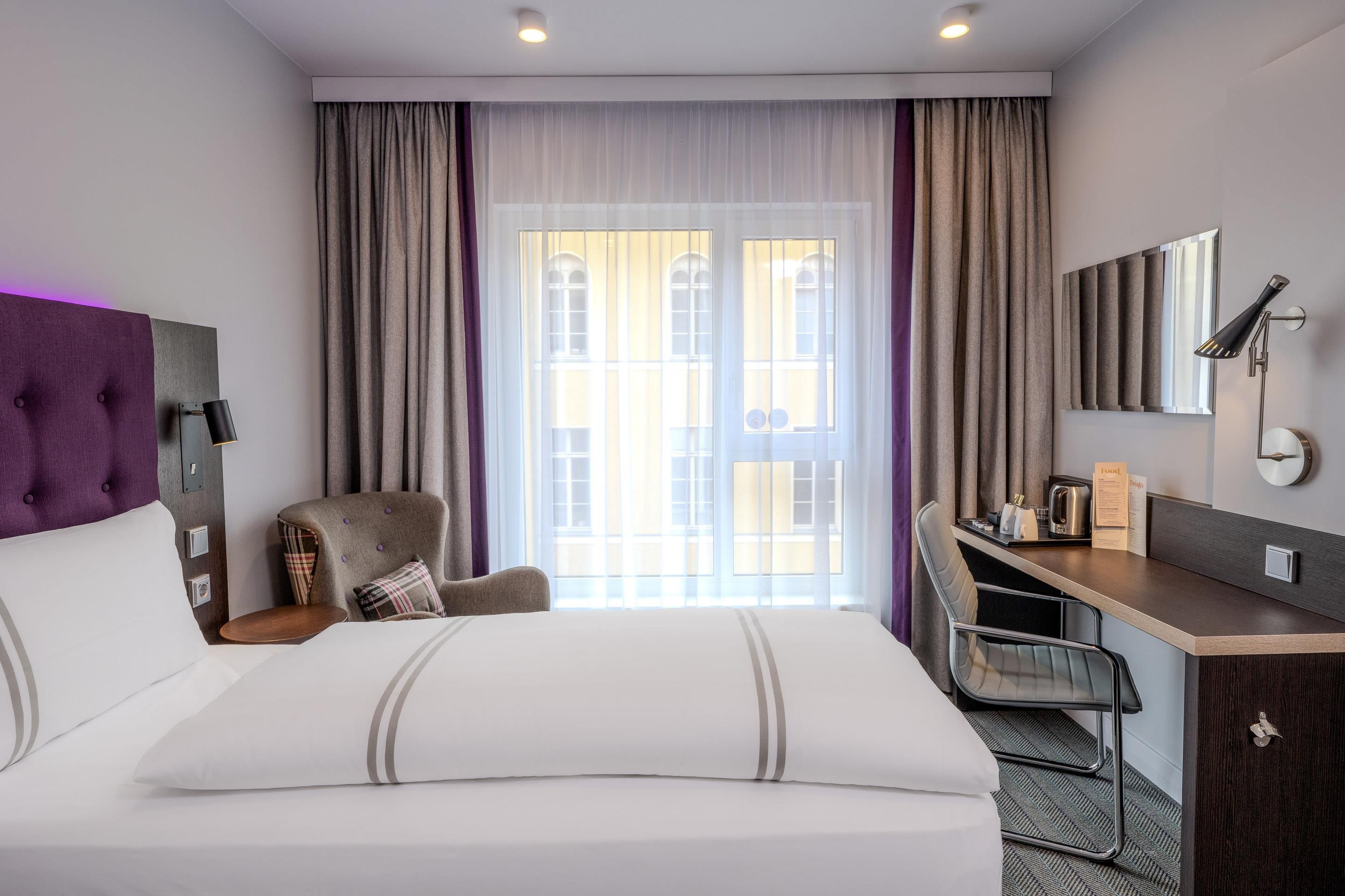 Une chambre d’hôtel comprenant un lit, un bureau, une chaise, une fenêtre avec des rideaux et une lampe fixée au mur.