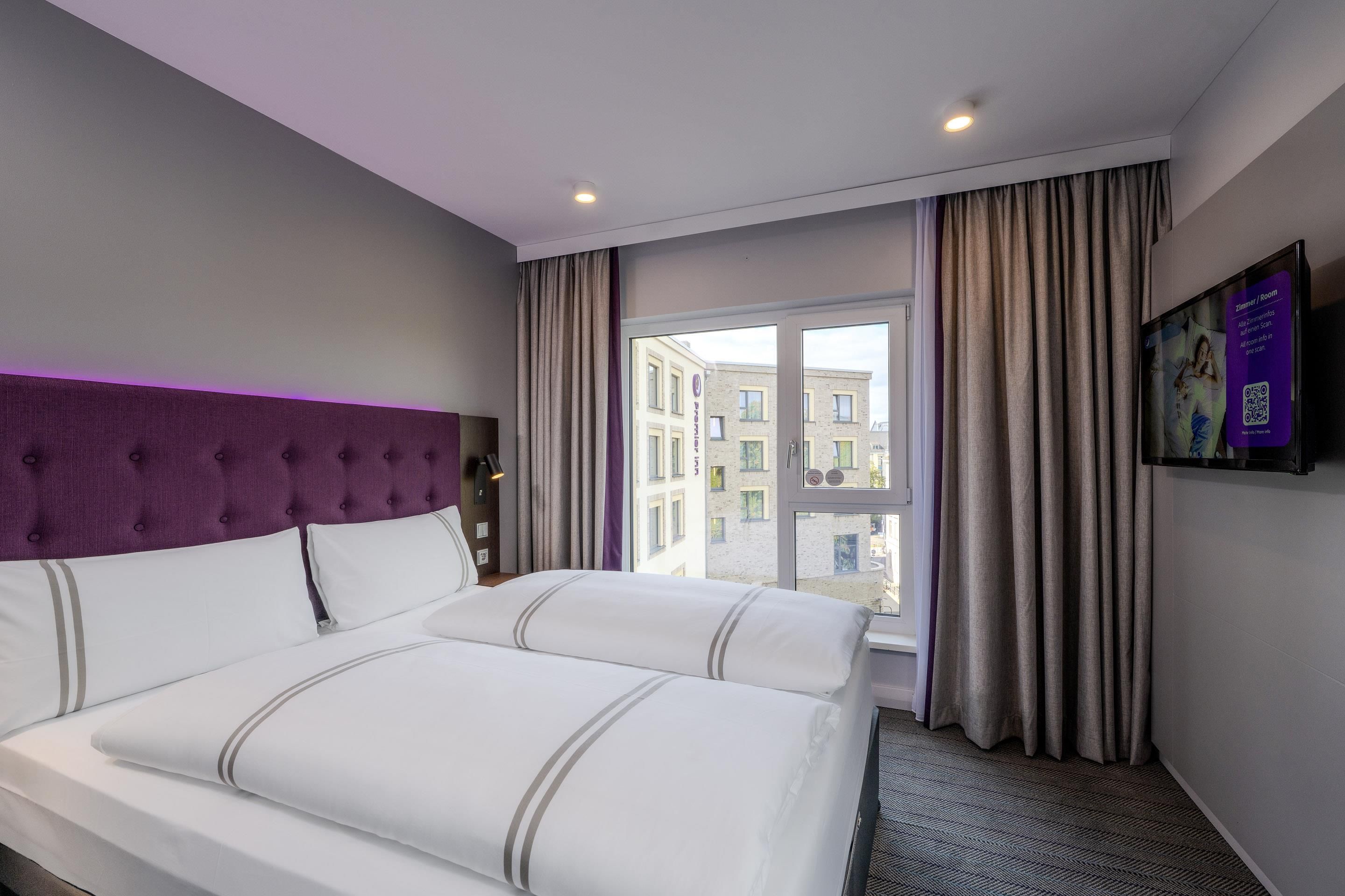 A hotel room with a bed, a headboard, a window with curtains, and a wall-mounted TV.
