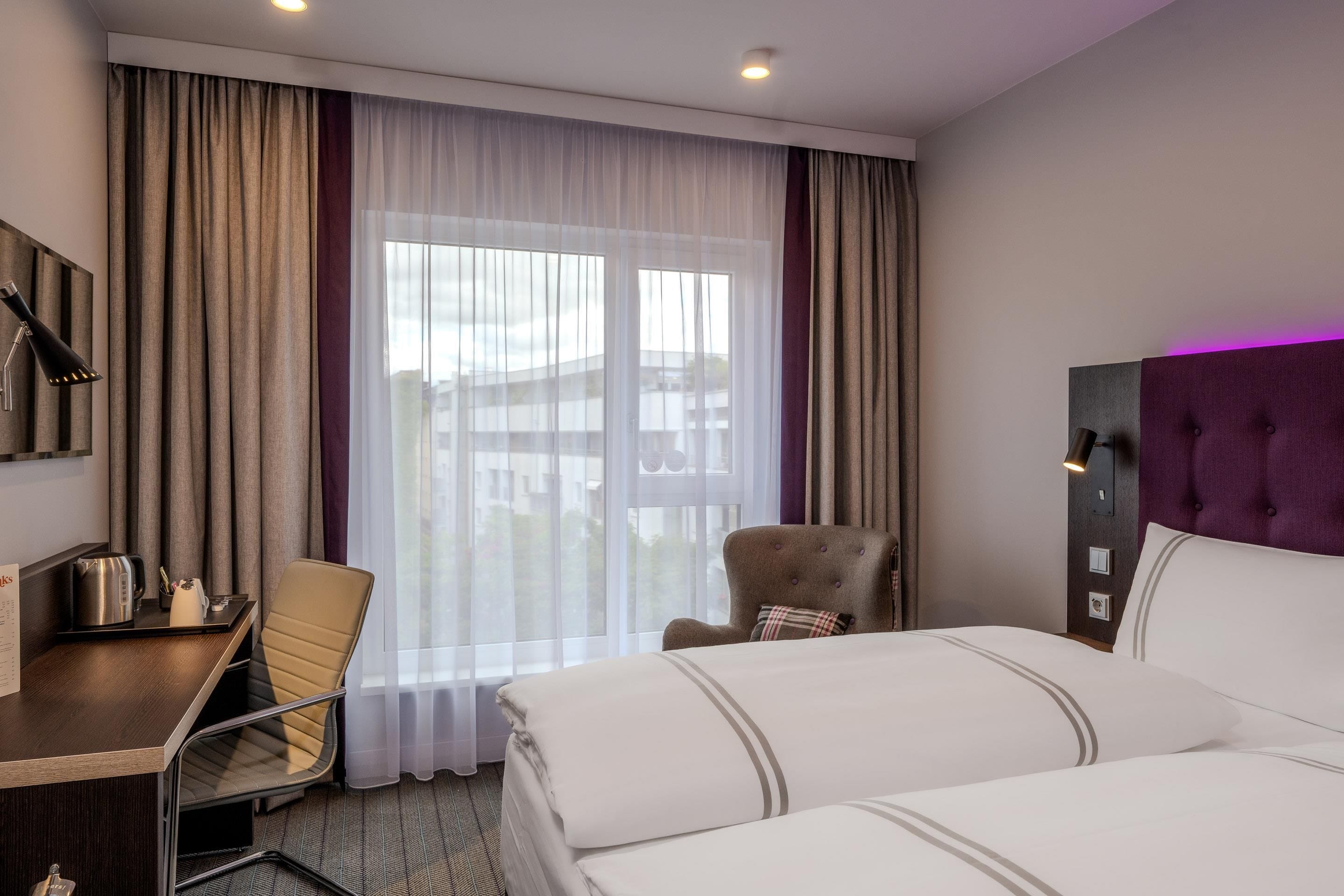 A hotel room with a bed, a desk, a chair, a teddy bear, and a window with curtains.