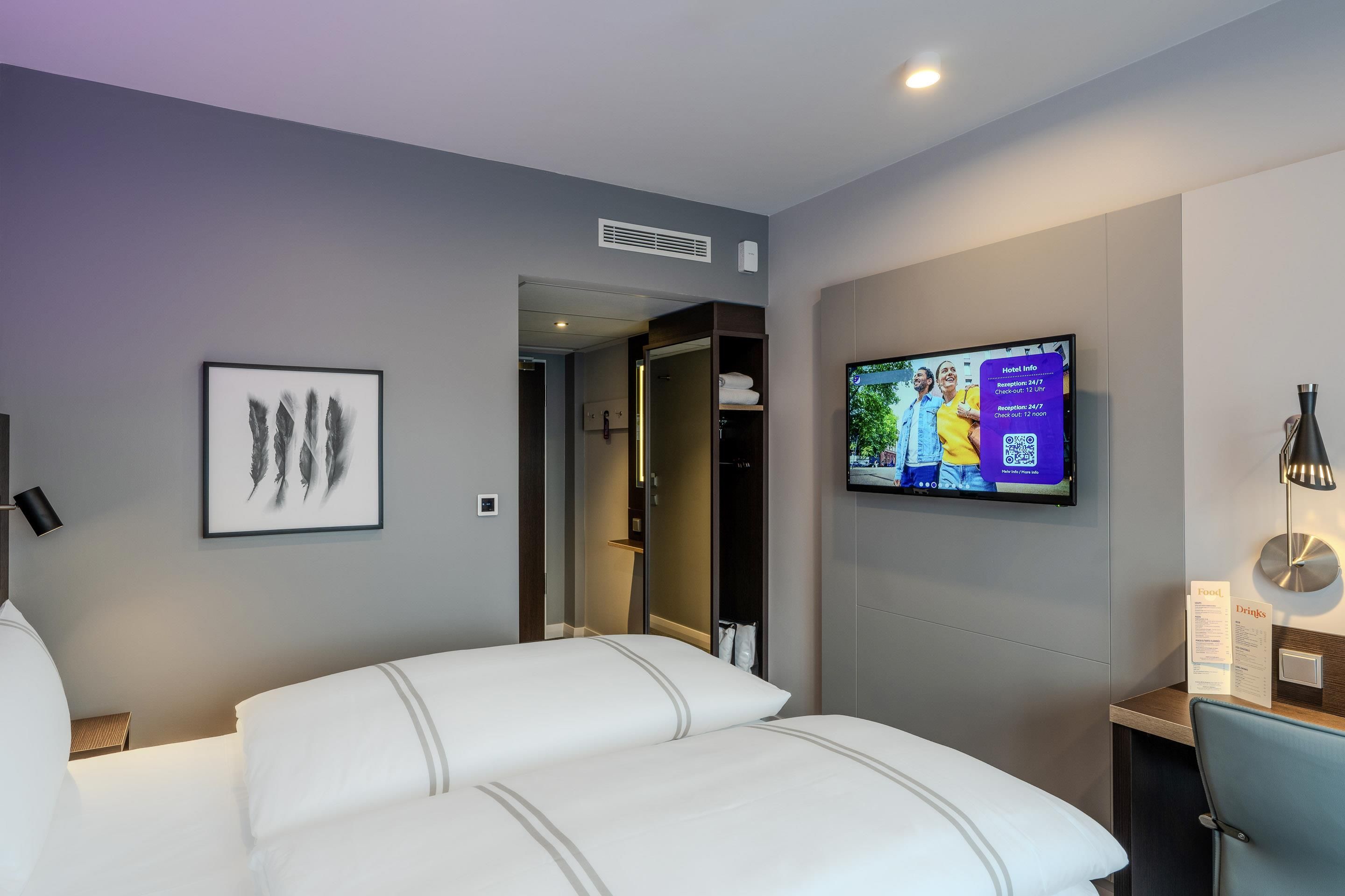 Una camera d'albergo con un letto, una televisione a muro che mostra informazioni dell'hotel e un'opera d'arte incorniciata.