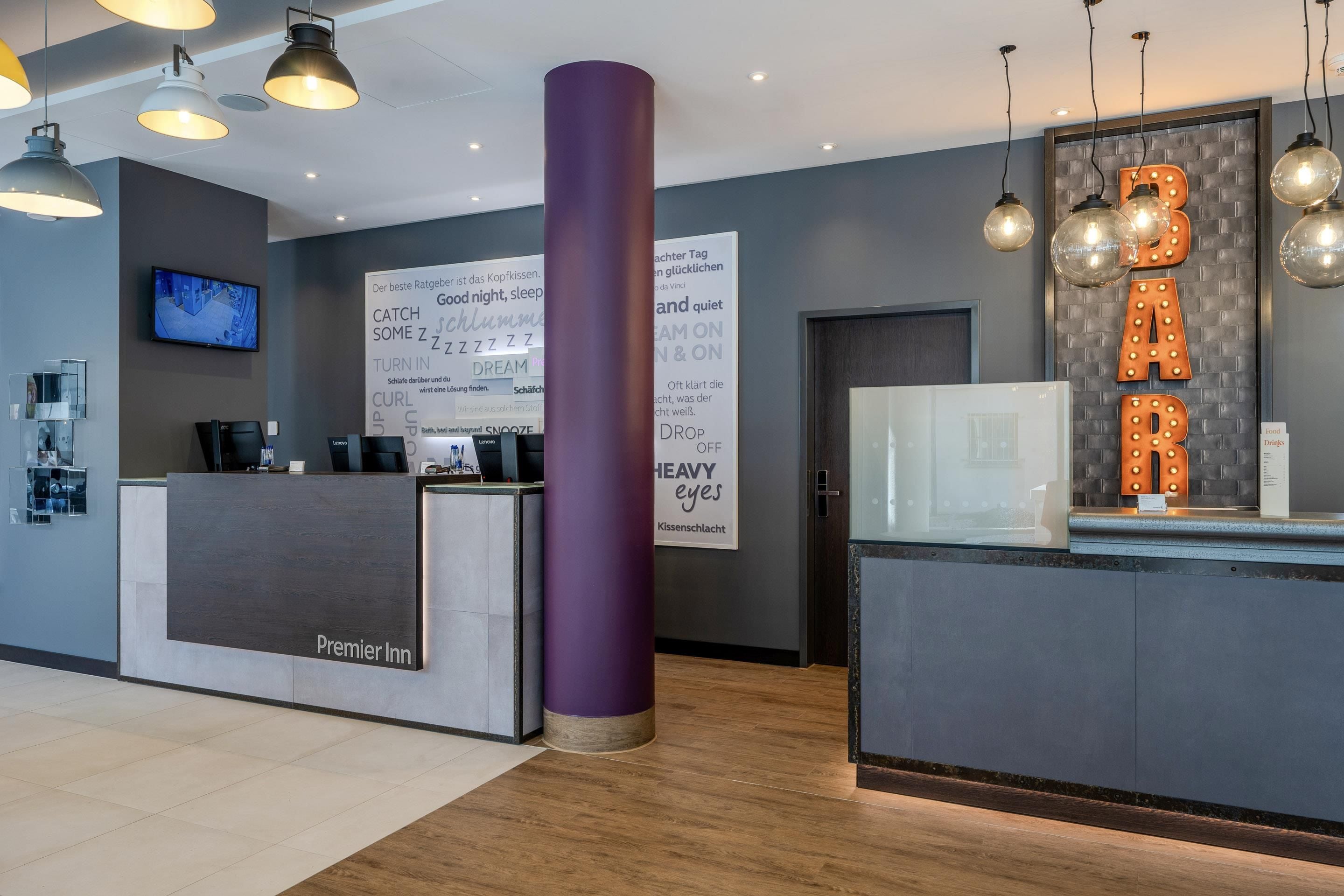 Ein Hotel-Empfangsbereich im modernen Stil, mit einer Rezeption mit der Beschriftung 'Premier Inn', einer großen violetten Säule und einem leuchtenden 'BAR'-Schild.