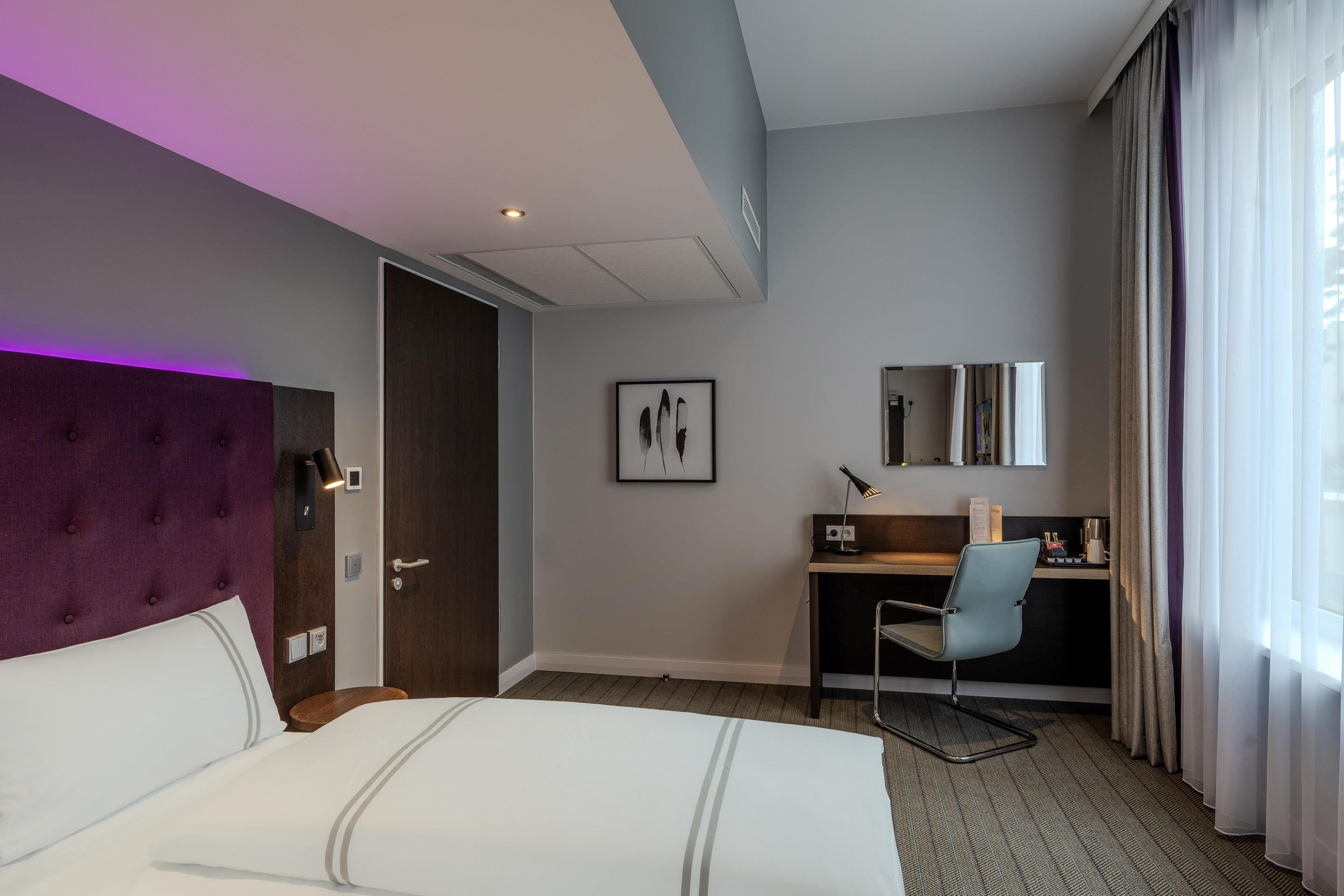 Habitación de hotel con cama, escritorio, silla, ventana con cortinas e imagen colgada en la pared.