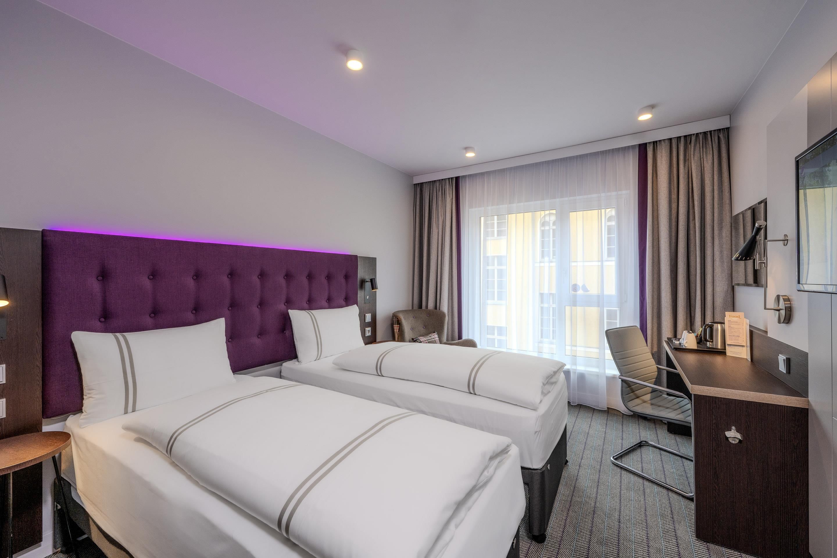 A hotel room with two beds, a desk, a chair, and a large window with curtains.