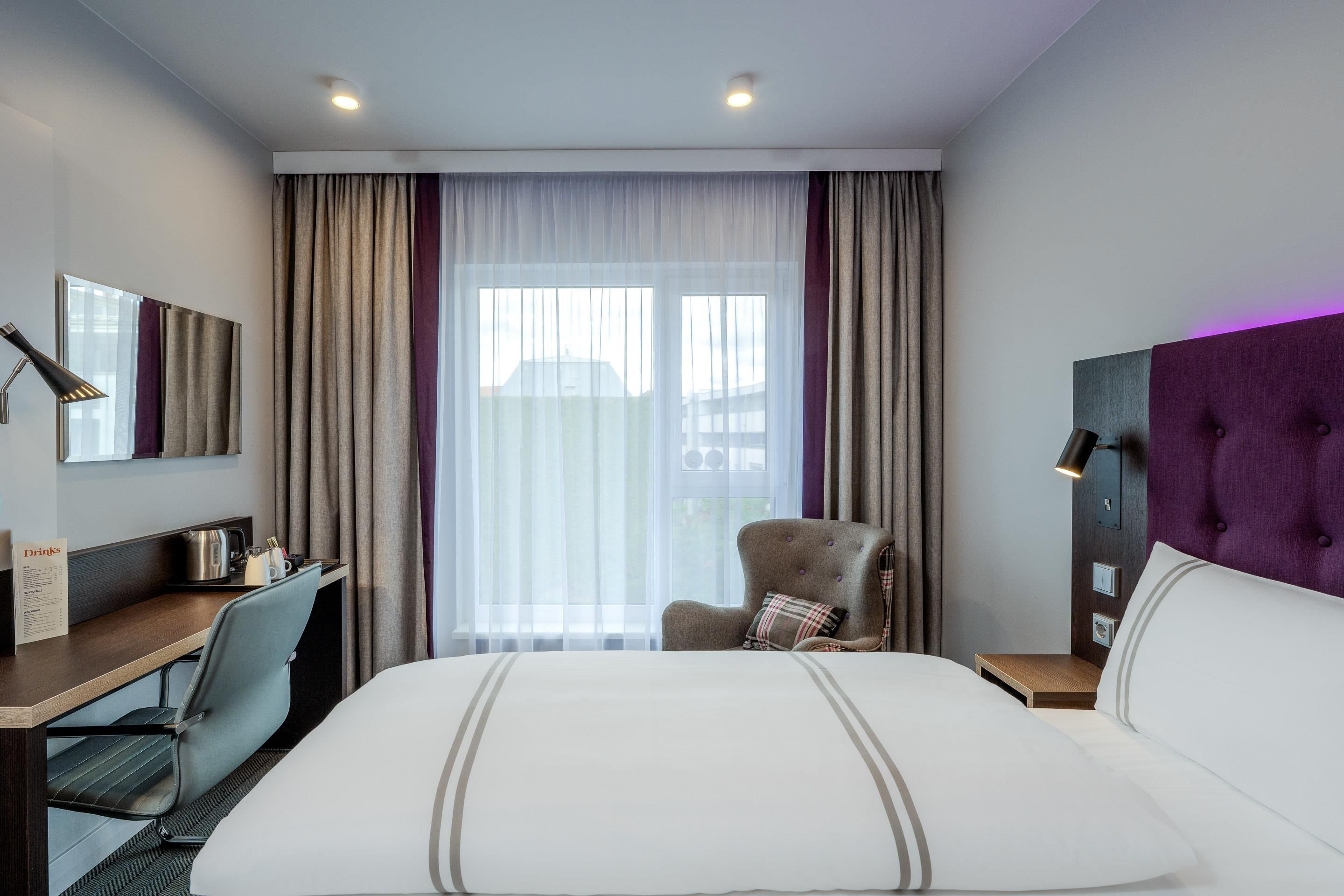 A hotel room with a bed, a desk, a chair, a nightstand, and a window with curtains.