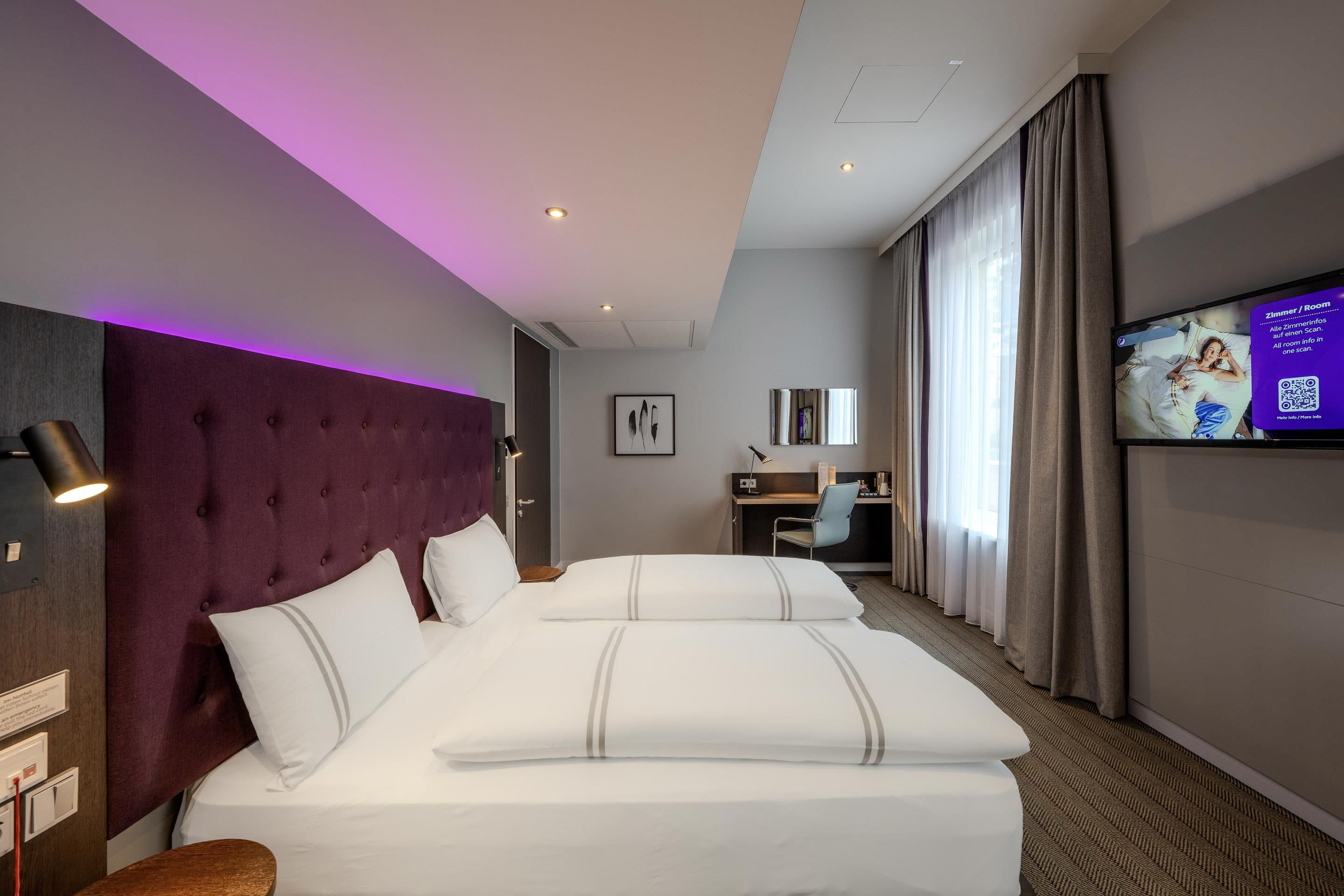 Ein Hotelzimmer mit einem großen Bett, einem Kopfteil, einem Schreibtisch, einem Stuhl, einem Fernseher und einem Vorhangfenster.