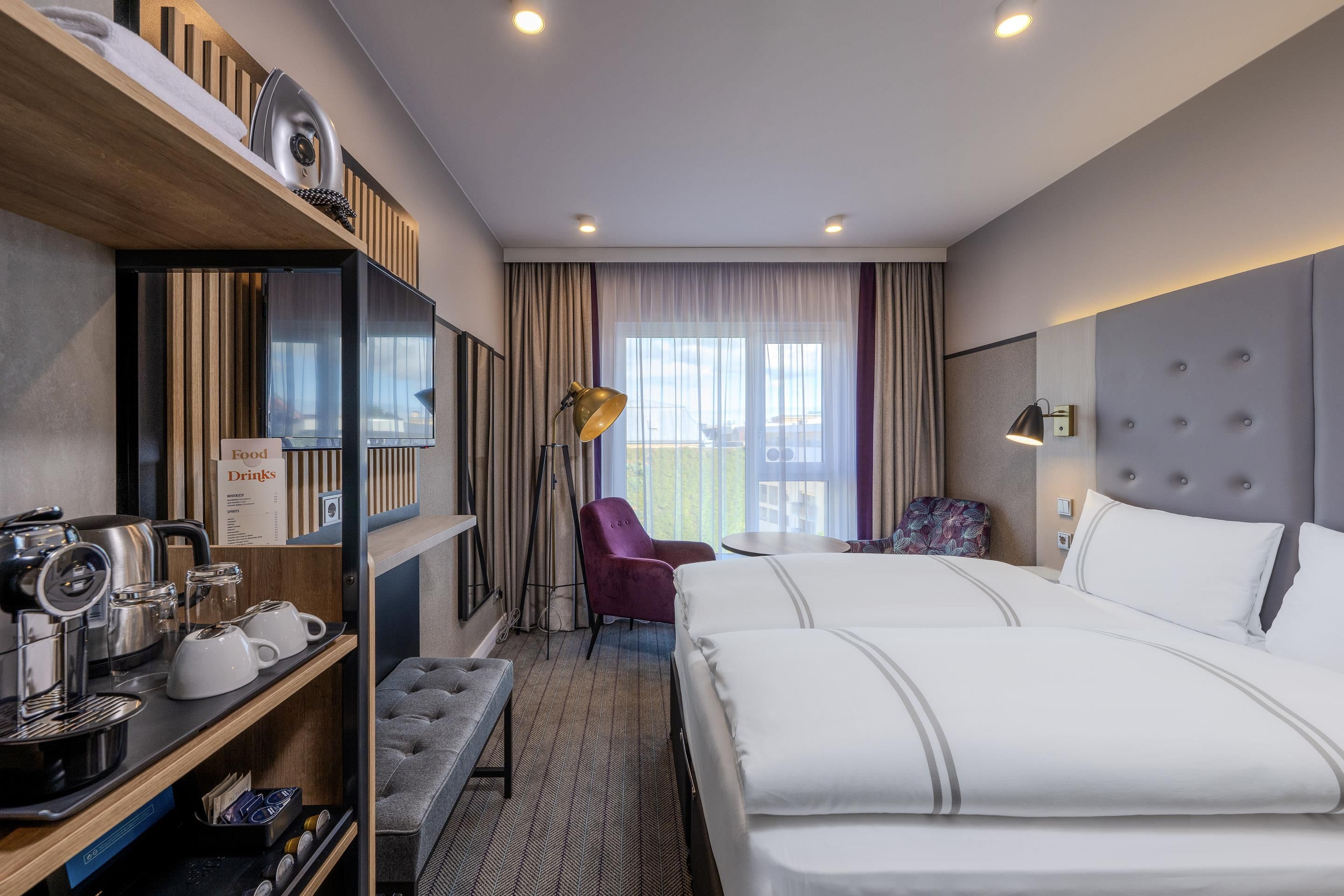 A hotel room with a bed, a seating area with a chair, a small table, a coffee machine, and a television.
