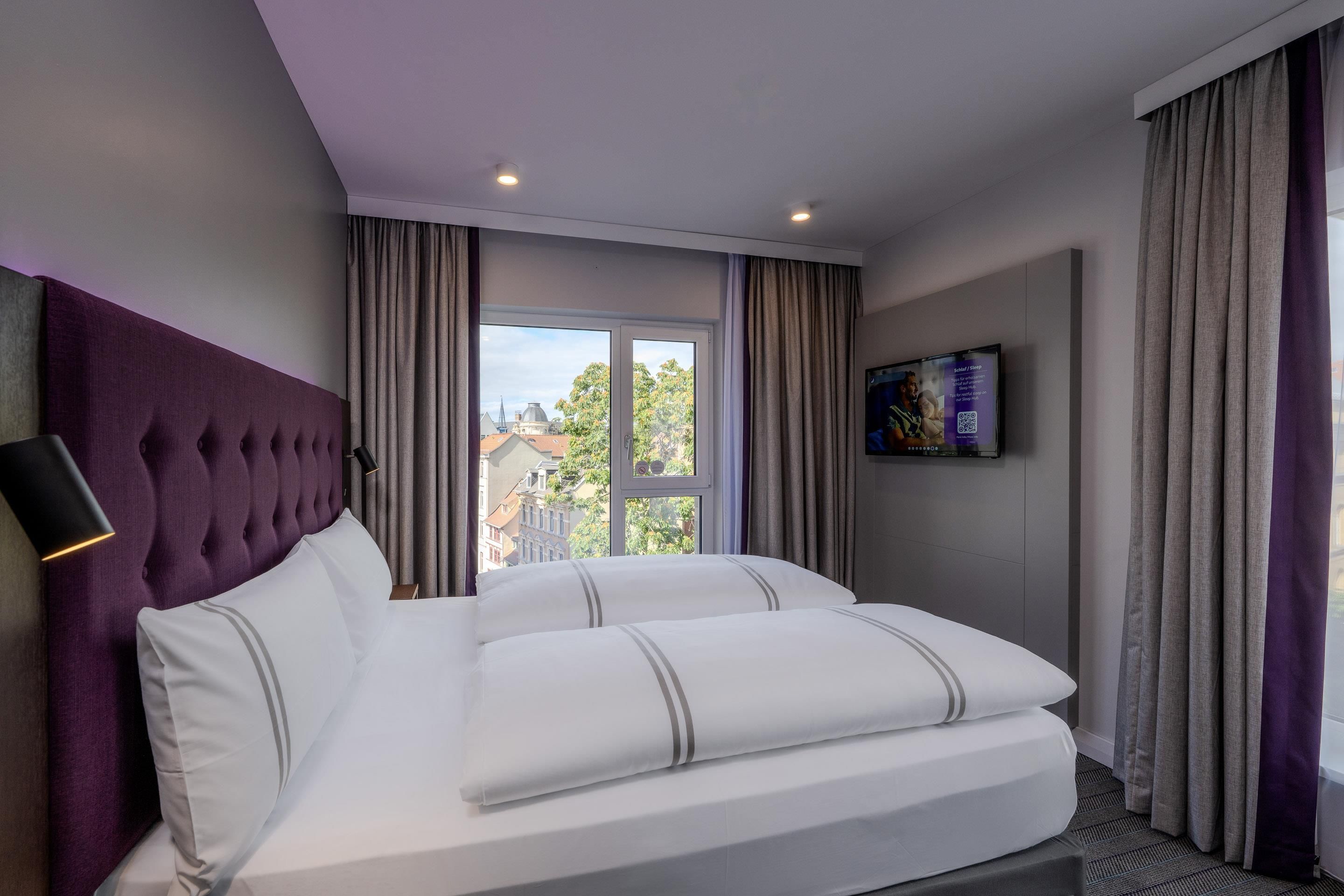 Ett hotellrum med en stor säng, en sänggavel, en tv och utsikt över byggnaderna utanför fönstret.