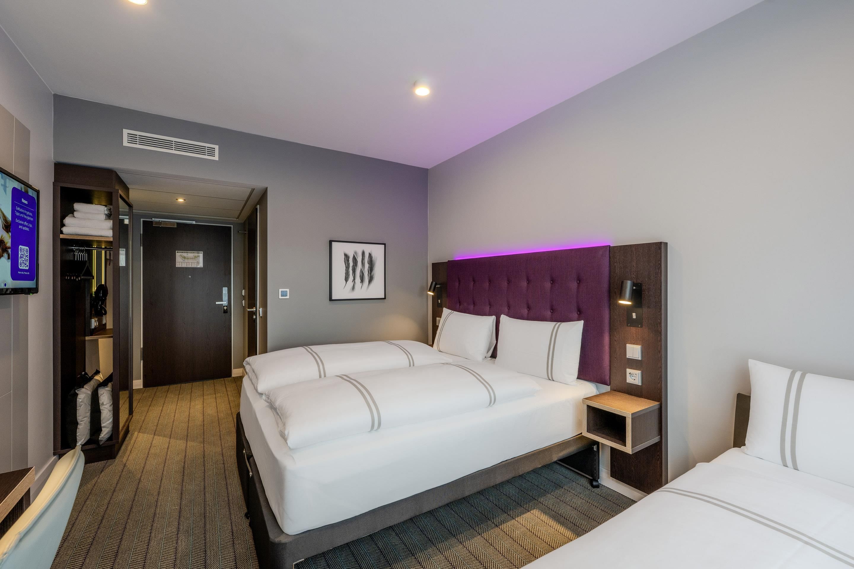 A hotel room with a large bed, a headboard with a purple accent, a small bedside table, and a wall-mounted TV.