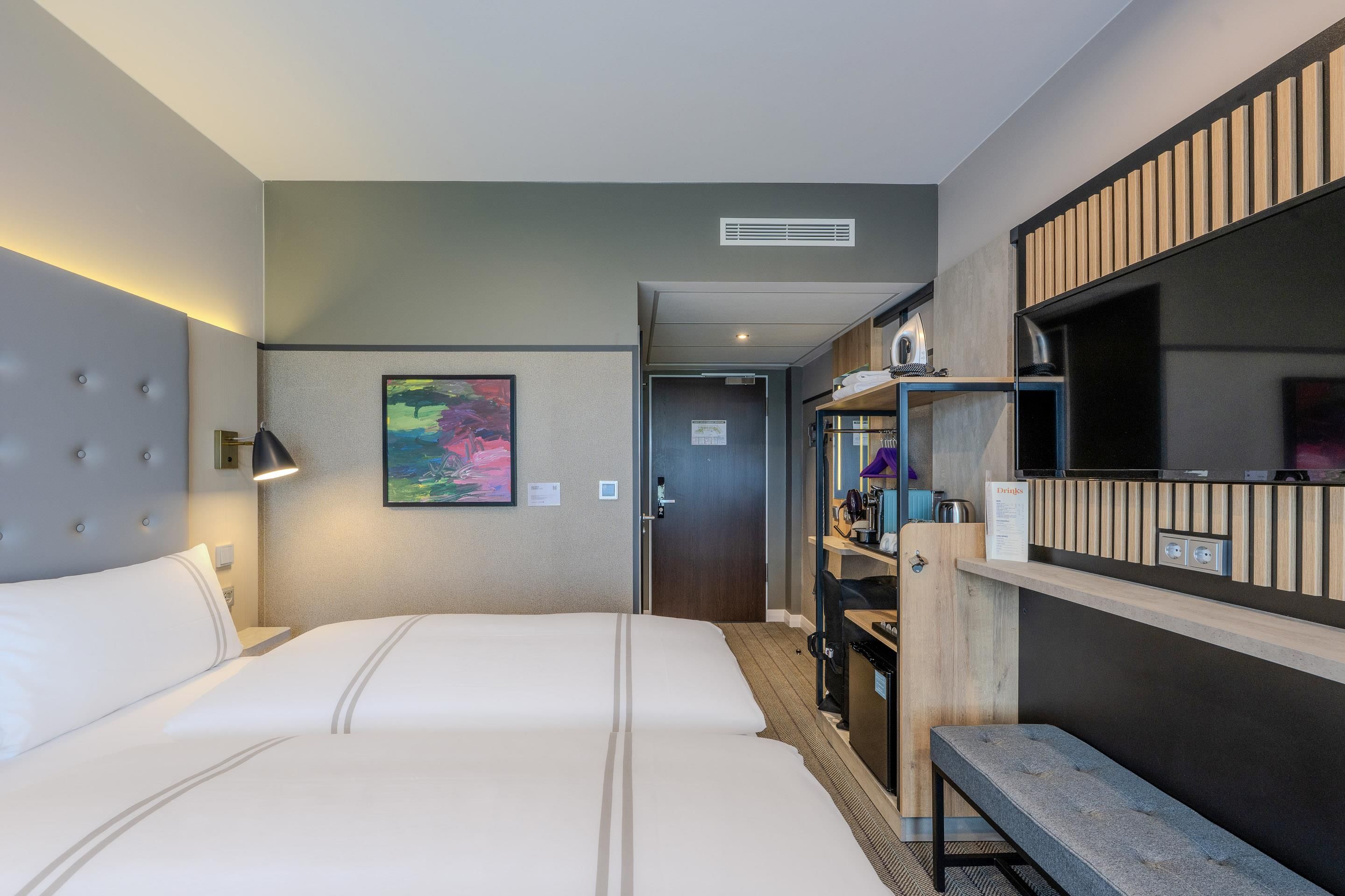 Habitación de hotel con una cama grande, un escritorio con computadora, un espejo y un cuadro en la pared.
