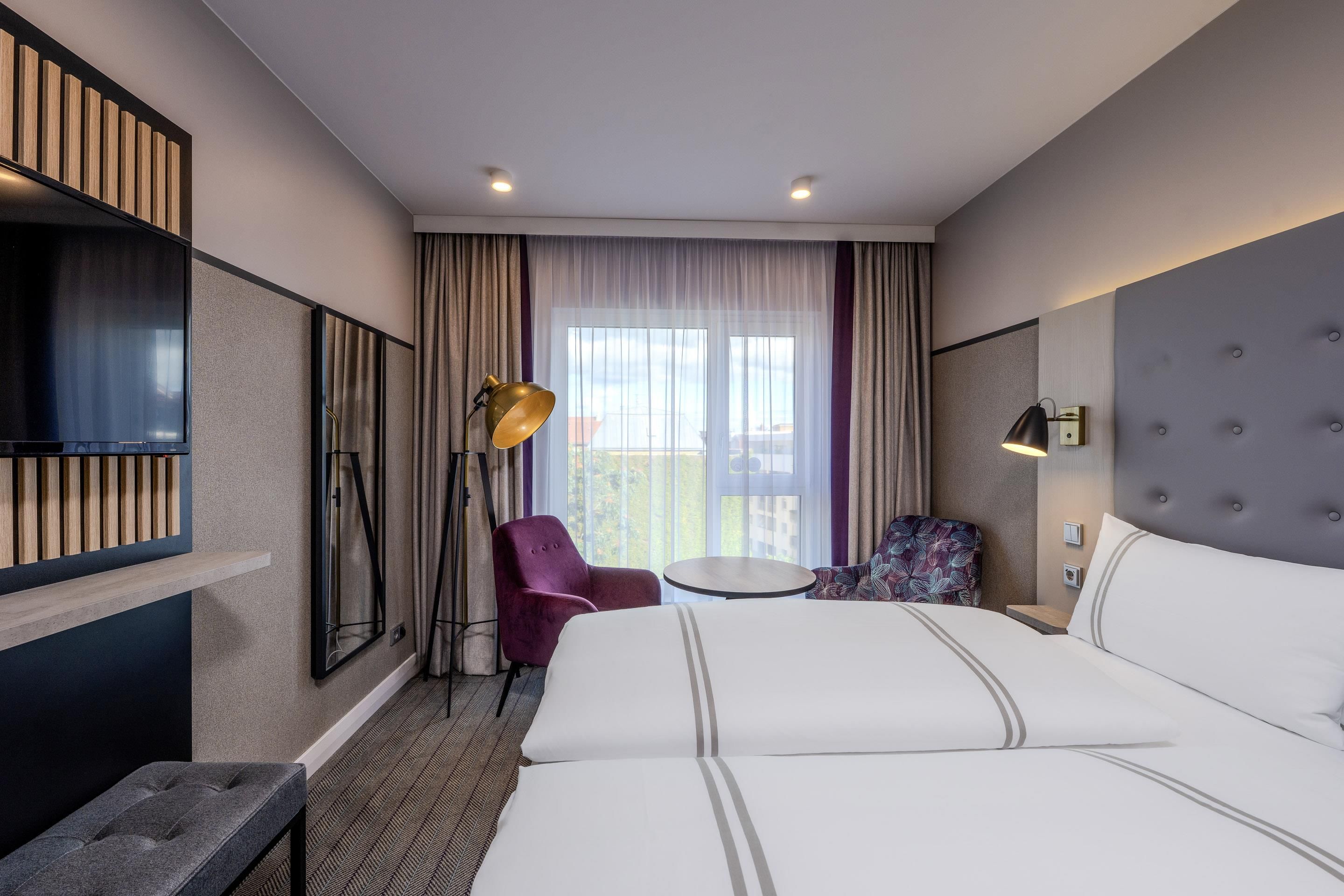 A hotel room with a large bed, a desk, a chair, a television, and a balcony with curtains.