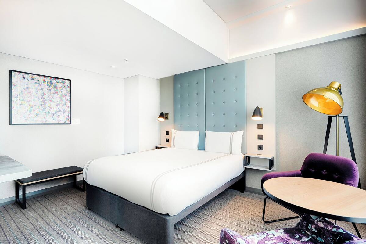 Moderni hotellihuone, jossa on suuri sänky, yöpöydät, penkki, pyöreä pöytä ja violetti tuoli.
