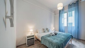 Une chambre à coucher comprenant un lit, deux tables de chevet, une commode, une fenêtre avec des rideaux et un luminaire suspendu.