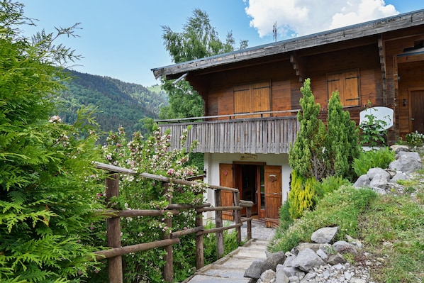 Un chalet en bois doté d’un balcon, entouré de verdure et avec une chaîne de montagnes en toile de fond.