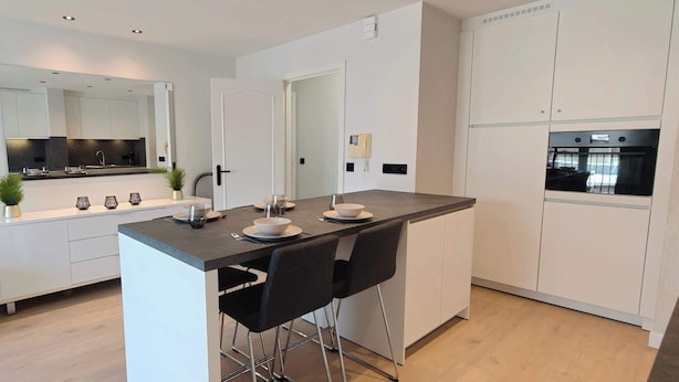 Una cocina moderna con una isla central, taburetes de bar, un sofá negro y un espejo grande.