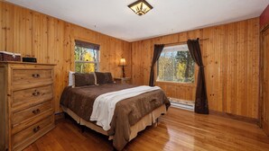 Un dormitorio con paneles de madera, una cama, un tocador, una lámpara de noche y ventanas.