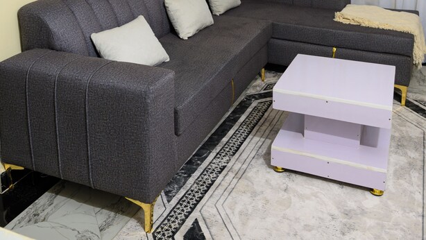 A living room with a grey sectional sofa, a white coffee table, a patterned rug, a painting on the wall, and a window with sheer curtains. - Lovely 2-bedroom apartment in beautiful Thika for a relaxing stay (Thika)
