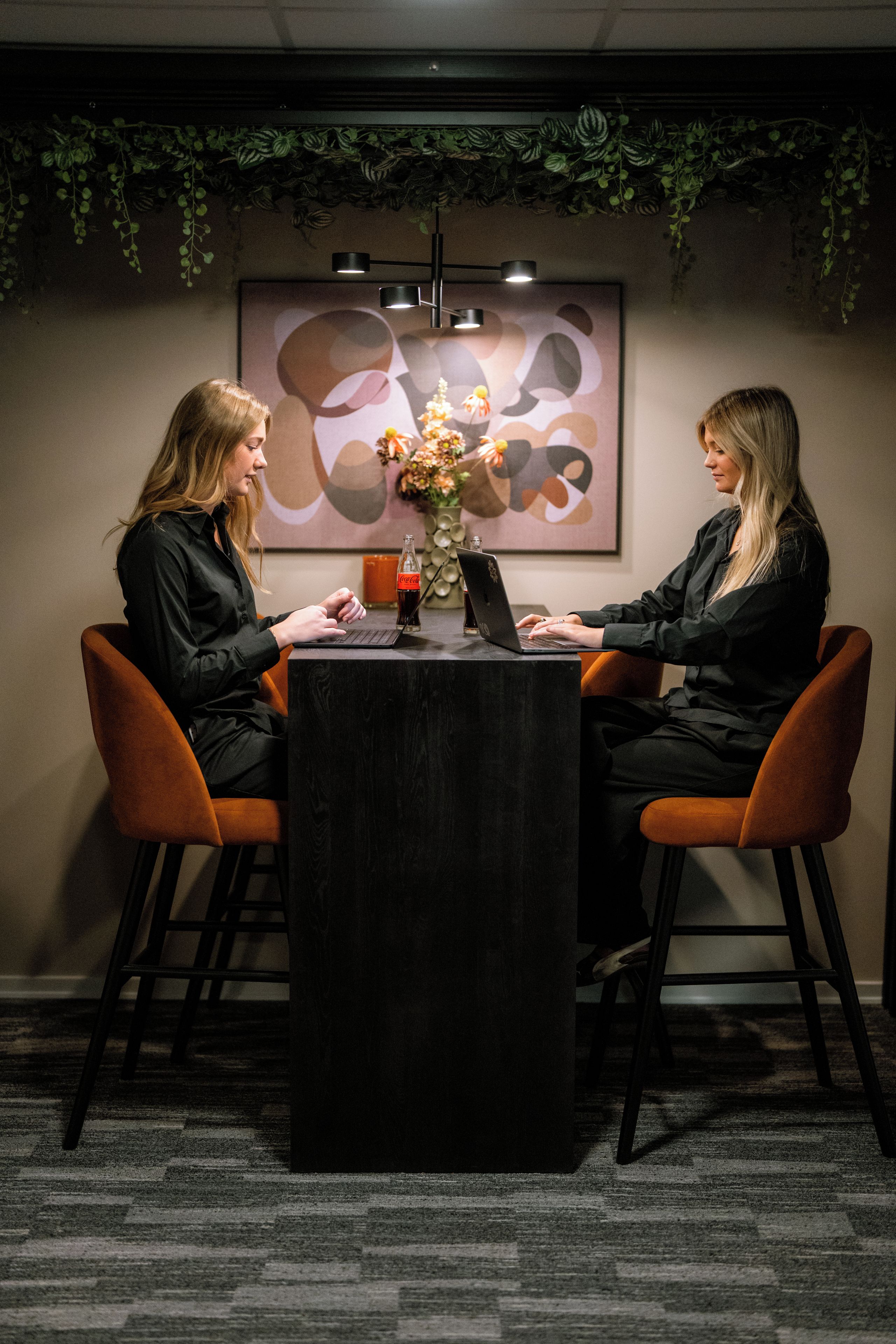 Twee personen zitten aan een tafel met laptops, in een kamer met een lijst met abstract kunstwerk aan de muur en een hangplant erboven.
