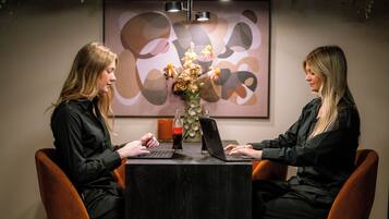 Twee personen zitten aan een tafel met laptops, in een kamer met een lijst met abstract kunstwerk aan de muur en een hangplant erboven.