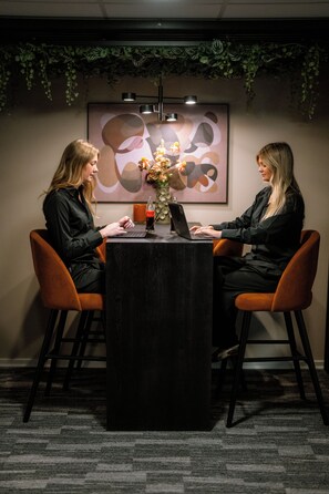 Dos personas sentadas en una mesa con computadoras portátiles, en una habitación con una obra de arte abstracta enmarcada en la pared y una planta colgando arriba.