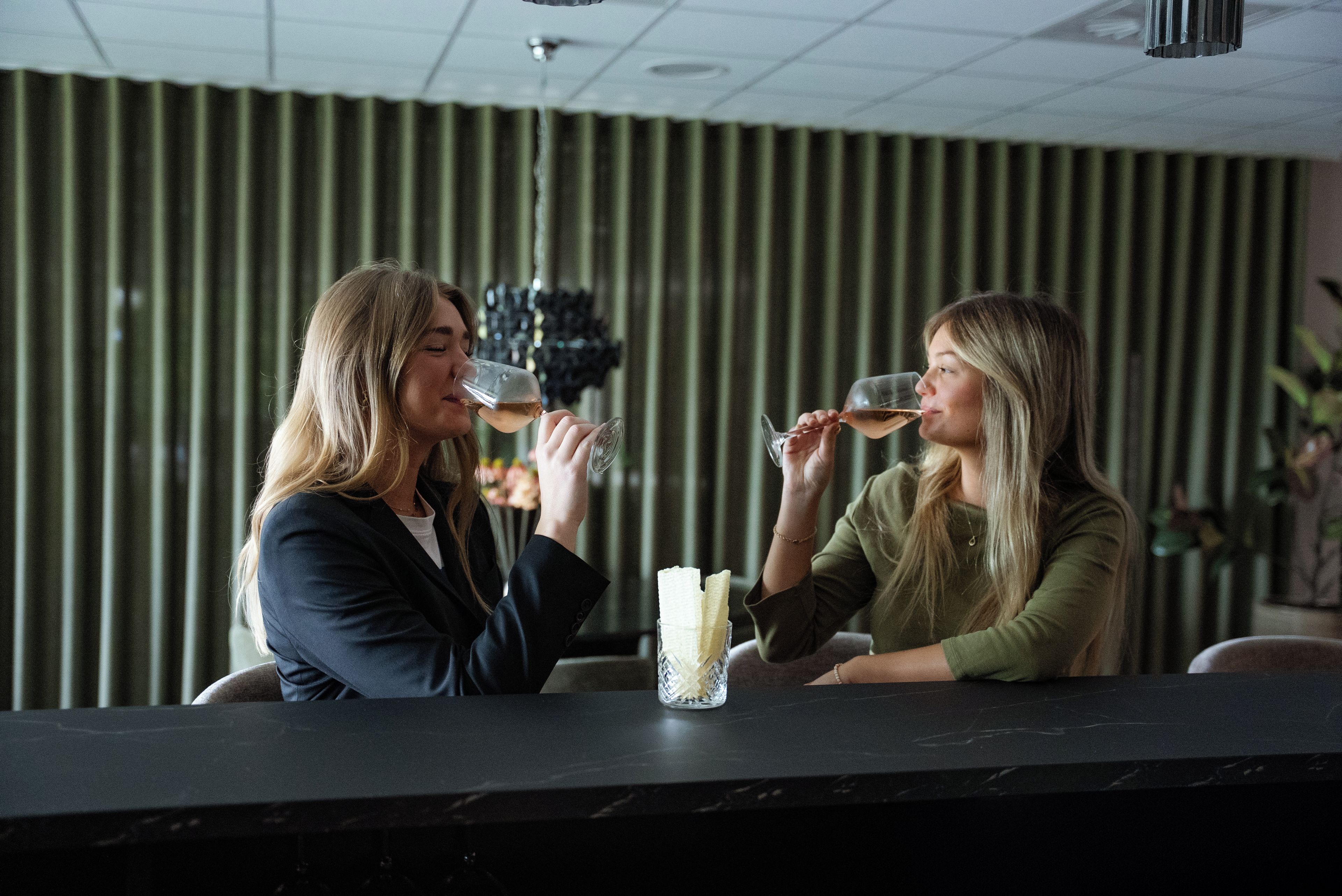 Two individuals seated at a bar with a glass of wine, a vase with flowers, and a striped wall in the background.