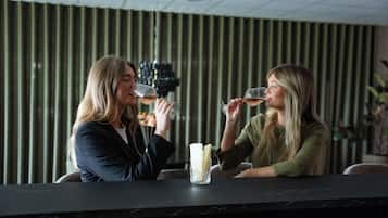 Two individuals seated at a bar with a glass of wine, a vase with flowers, and a striped wall in the background.