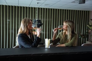 Dos personas sentadas en un bar, con una copa de vino, un florero y una pared a rayas al fondo.