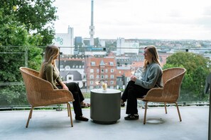 Dos personas sentadas en sillas de mimbre alrededor de una mesa redonda, con una copa de vino, con vistas a un paisaje urbano con edificios y vegetación.