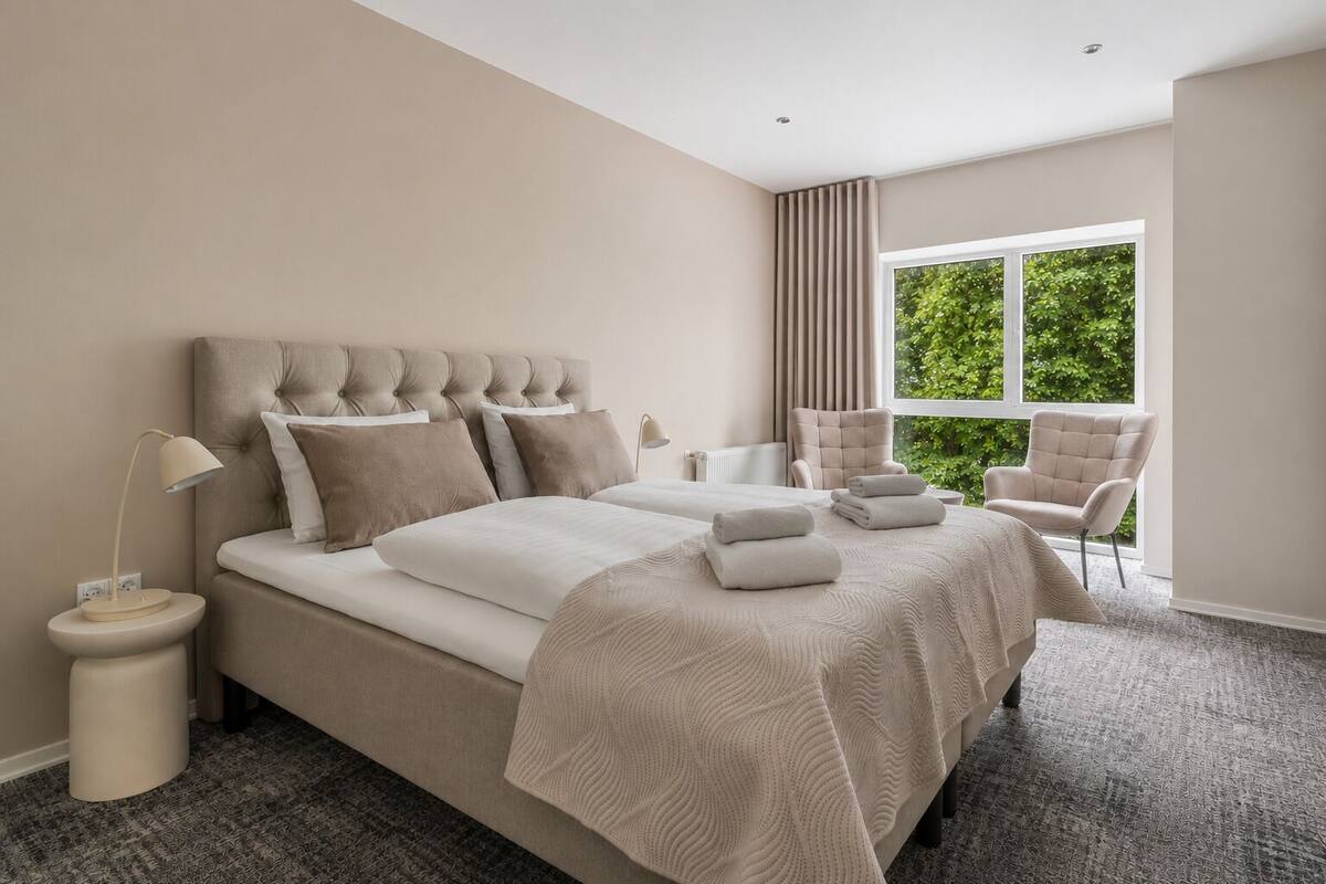 Un dormitorio con una cama grande, mesitas de noche, una silla y una ventana con vistas a zonas verdes.