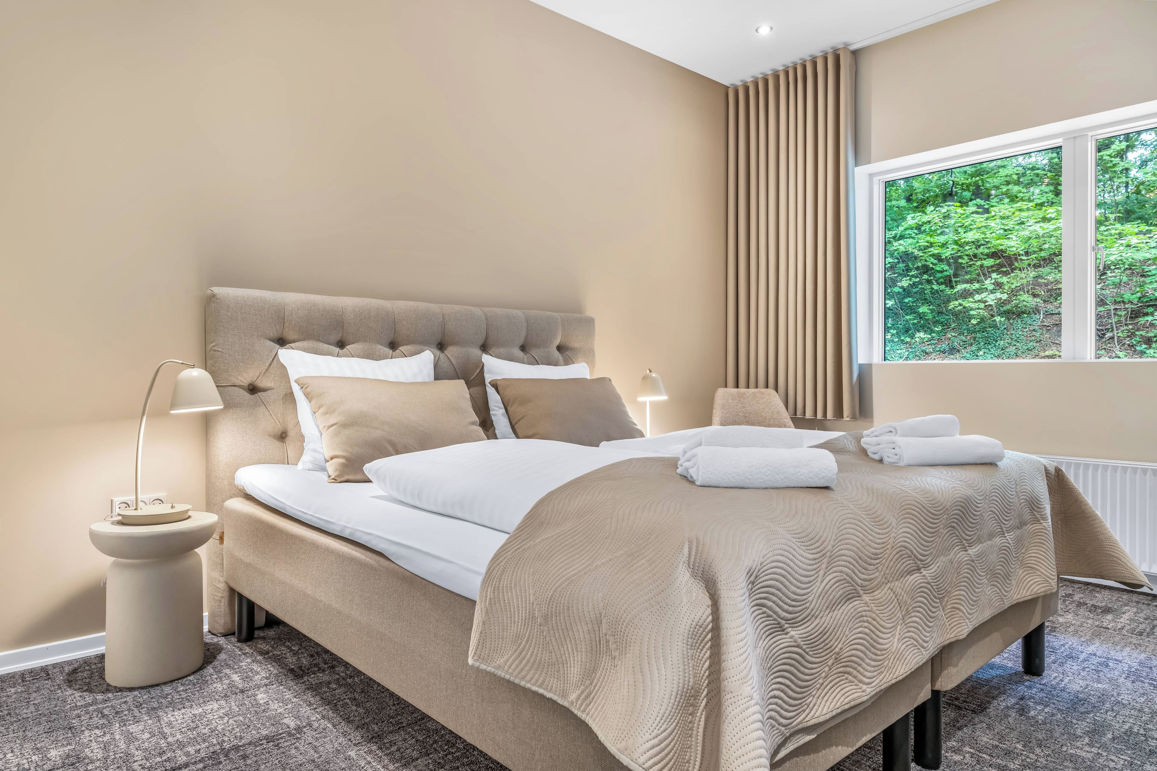Een slaapkamer met een groot bed, twee bedlampen, een raam met gordijnen en uitzicht op groen.