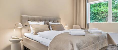 A bedroom with a large bed, two bedside lamps, a window with curtains, and a view of greenery outside.