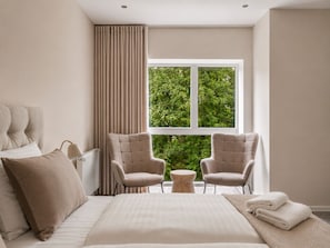 Un dormitorio moderno con una cama grande, dos sillones, una mesita redonda y un ventanal con vistas a zonas verdes.