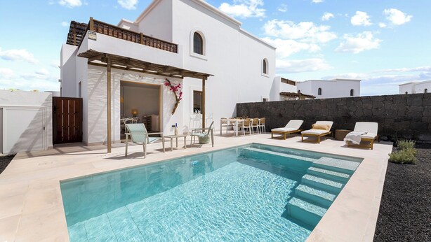 Una villa blanca moderna con piscina, mobiliario exterior y un muro de piedra.