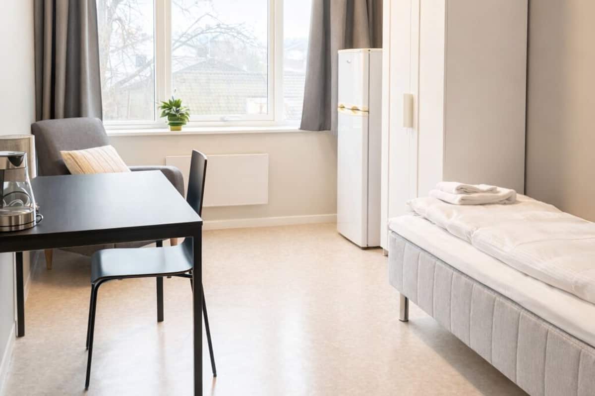 A compact room with a bed, a table with chairs, a radiator, a window with curtains, and a white wardrobe.