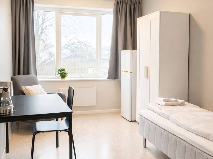 Una habitación pequeña con cama, una mesa y sillas, un radiador, un ventanal con cortinas y un armario blanco.