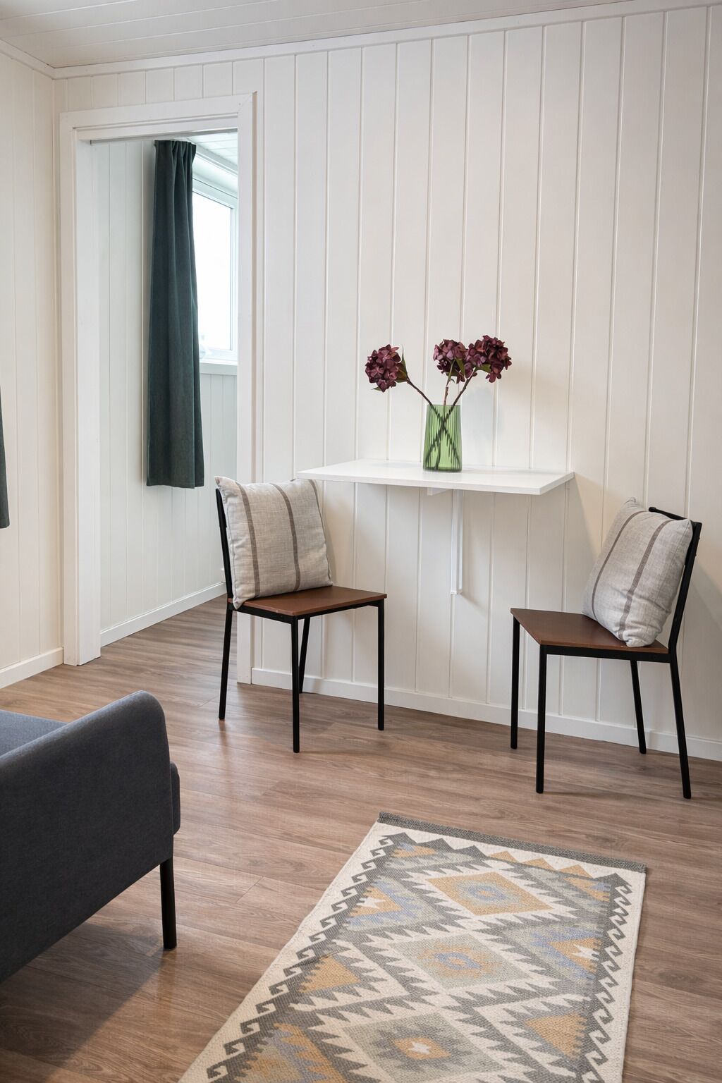 A room with a white paneled wall, a small table with a vase of flowers, two chairs, a patterned rug, and a dark sofa.