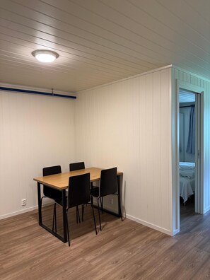 A room with a wooden dining table, four chairs, a ceiling light, and a doorway leading to a bedroom.