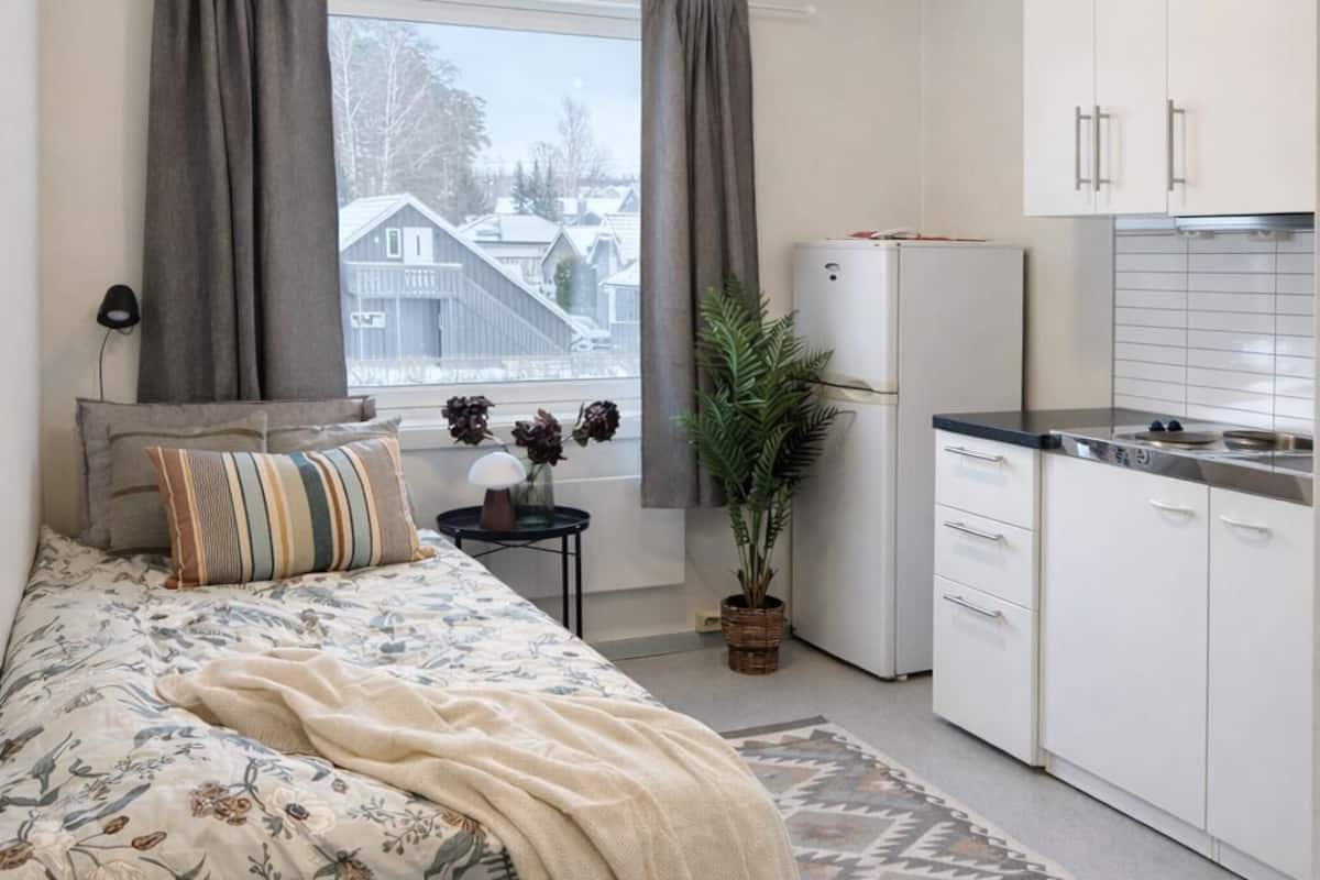 A bedroom with a bed, a bedside table, a lamp, a window with curtains, a potted plant, and a kitchen area with cabinets and a refrigerator.