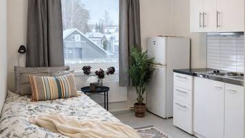 A bedroom with a bed, a bedside table, a lamp, a window with curtains, a potted plant, and a kitchen area with cabinets and a refrigerator.