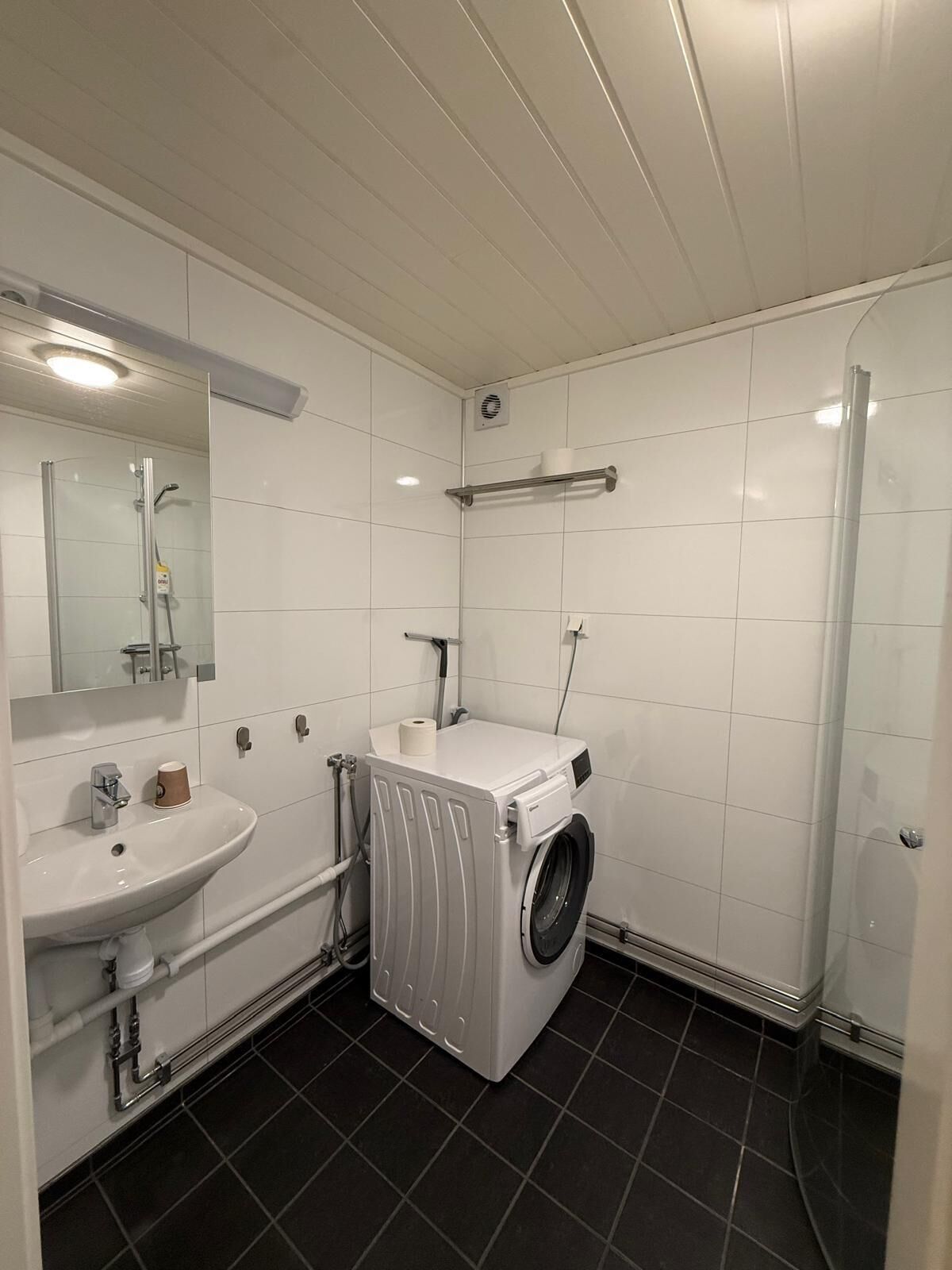 Un baño moderno con lavamanos, espejo, azulejos blancos, lavadora y zona de ducha.