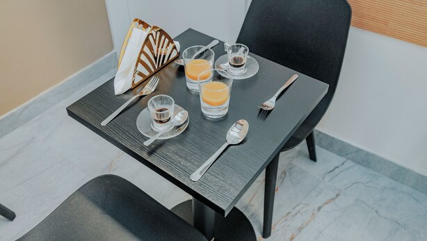 Uma mesa de jantar posta com garfo, colher, dois copos de suco de laranja, uma xícara pequena de café e um objeto decorativo.