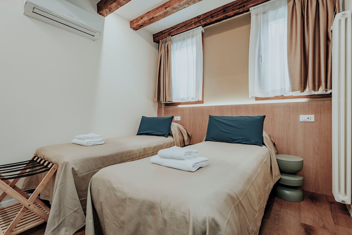 A hotel room with two beds, a wooden chair, a radiator, and a window with curtains.