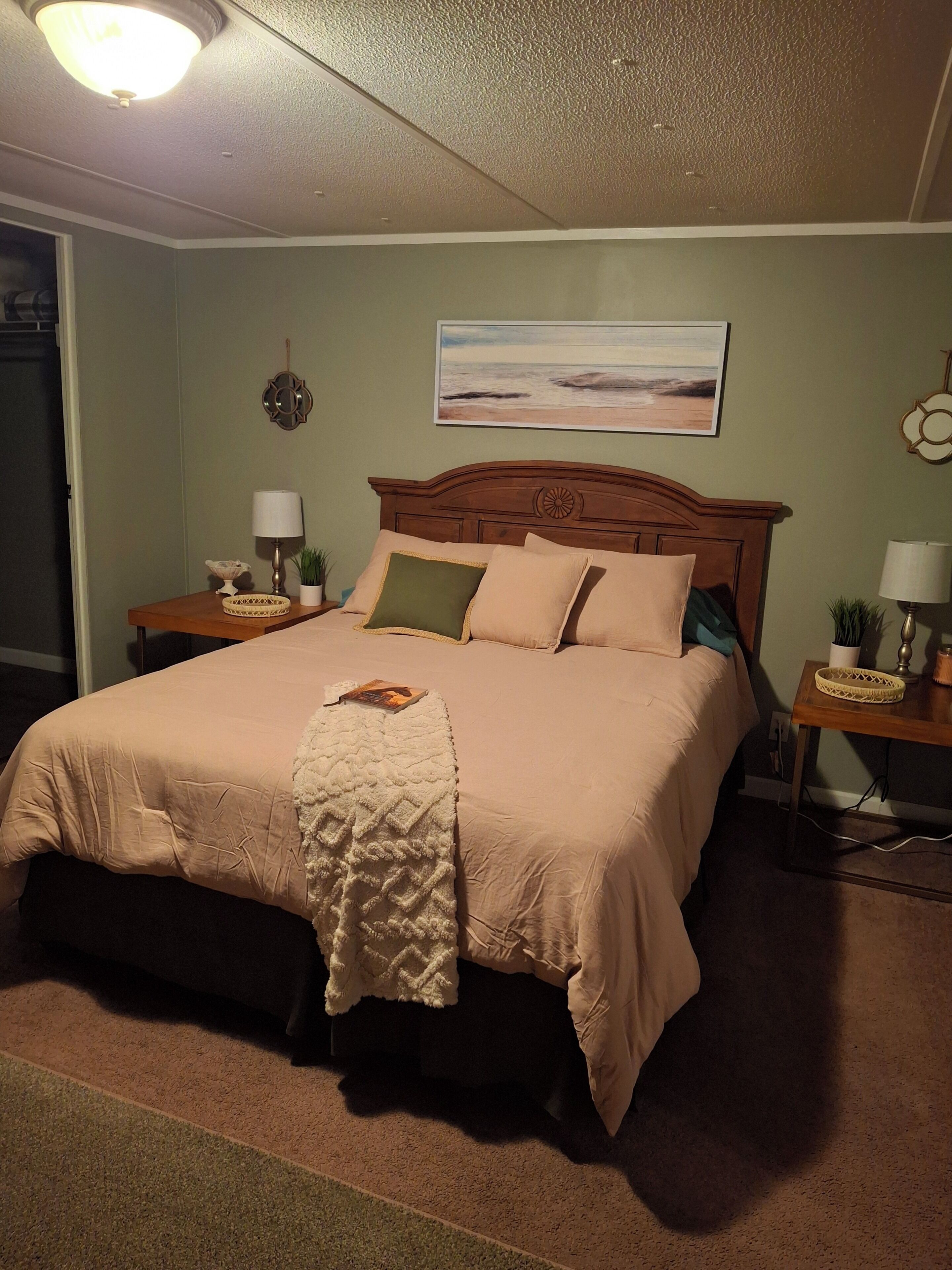 A neatly made bed with a headboard, two bedside tables with lamps, a wall clock, and a painting of a beach scene.