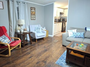 Ein Wohnzimmer mit Holzboden, einem roten Sessel, einem grauen Sofa, einem Couchtisch und einer Küche im Hintergrund.