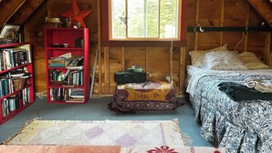 A cozy attic bedroom with a sloped ceiling, a bed with patterned bedding, a red bookshelf, and a window with a view of greenery. - 1-Bedroom Off Grid Cabin on Remote Island Across from Acadia National Park (Tremont)