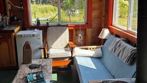 A cozy cabin interior with a wooden interior, a bed, a table with books, a chair, and a window with a view of greenery. - 1-Bedroom Off Grid Cabin on Remote Island Across from Acadia National Park (Tremont)