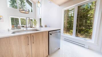 Une cuisine moderne équipée d’un lave-vaisselle, dotée de grandes fenêtres et offrant une vue sur une forêt.