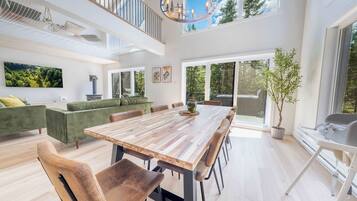 A spacious dining area with a long wooden table and modern chairs, a green sofa set, and a large window offering a view of the outdoors.