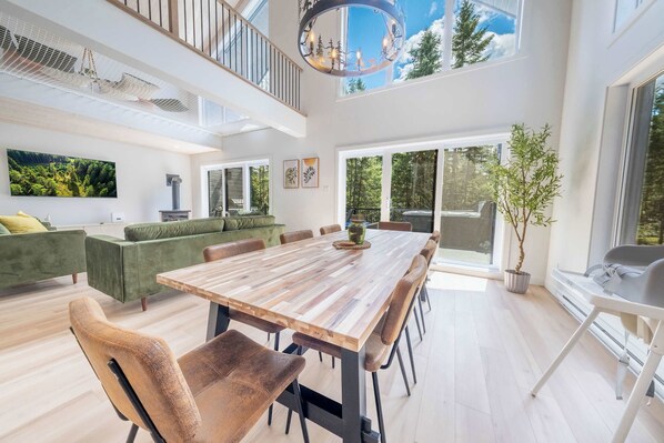 A spacious dining area with a long wooden table and modern chairs, a green sofa set, and a large window offering a view of the outdoors.