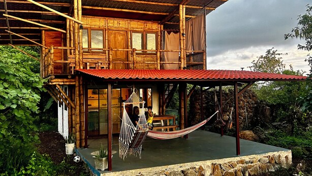 Una casa de madera de dos pisos con un balcón, una hamaca y un muro de piedra.