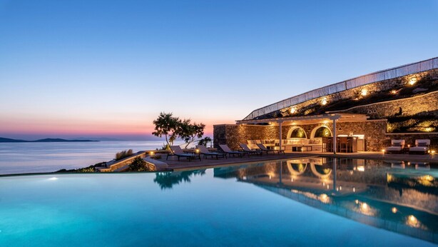A poolside area with lounge chairs, a stone building with arches, and a view of the sea. - Luxury Villa Roshell (Mykonos)